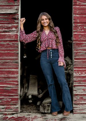 Woman in pink floral top and flared jeans poses in front of weathered red barn door in rustic outdoor photoshoot.