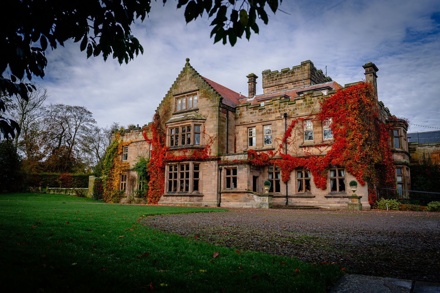 Panoramic view of an elegant stone mansion with fall foliage and manicured grounds. Ellingham Hall