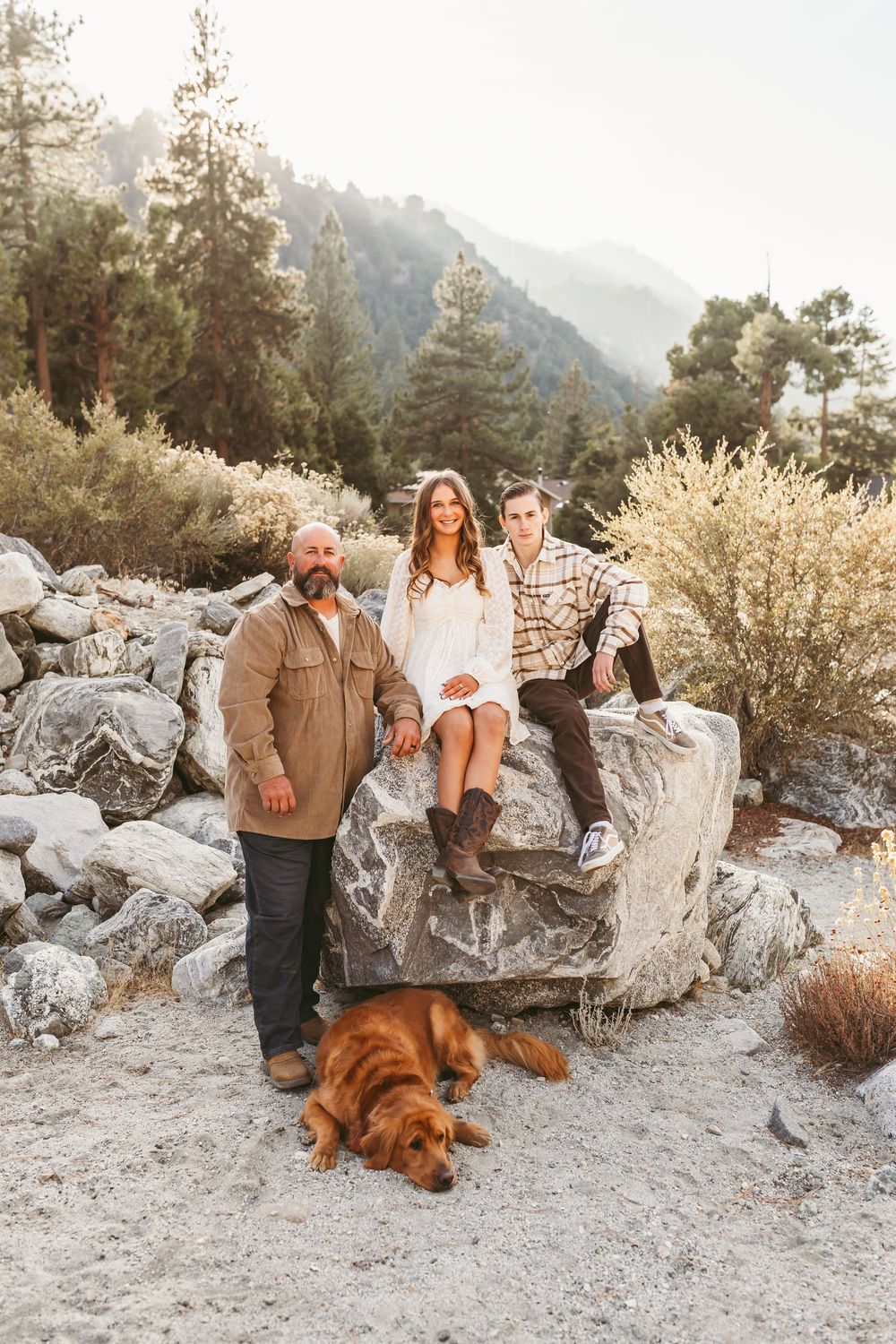 People and a golden retriever dog gather on rocky terrain with mountains and pine trees in the background.