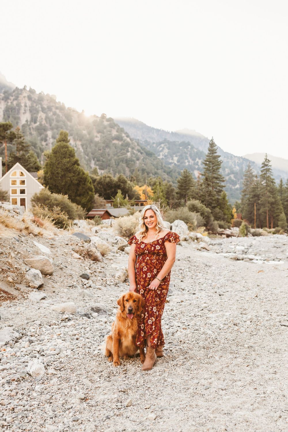 A Golden Retriever sits with someone in a floral dress against a mountainous backdrop with pine trees.