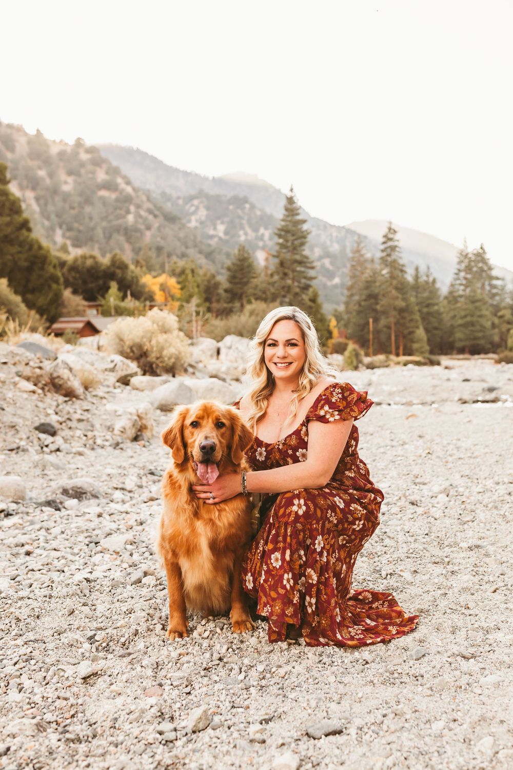 A golden retriever and person in a floral dress sit together on rocky ground with mountains in the background.