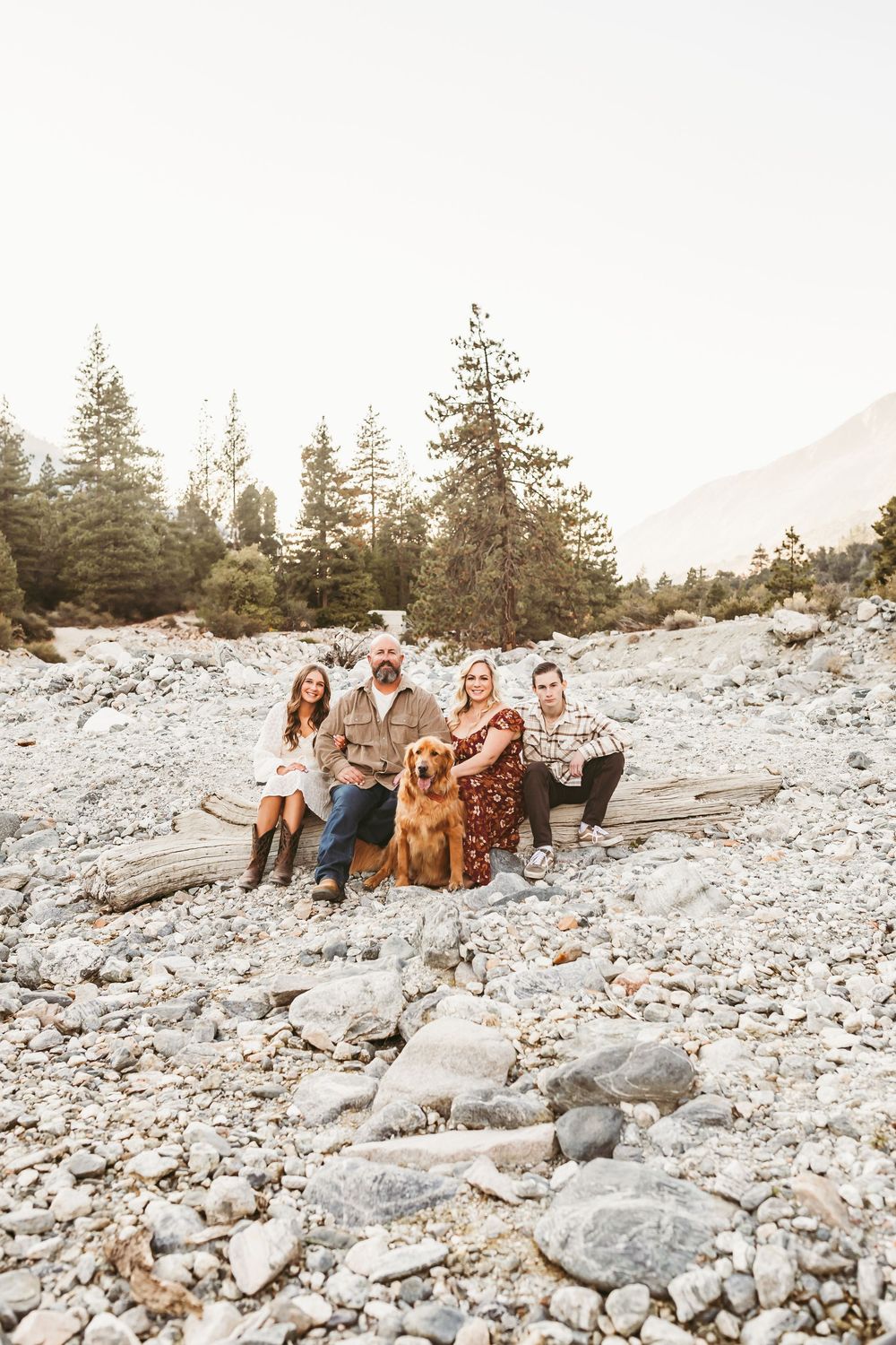 A group relaxes on rocky terrain with a Golden Retriever against a mountain landscape at sunset.