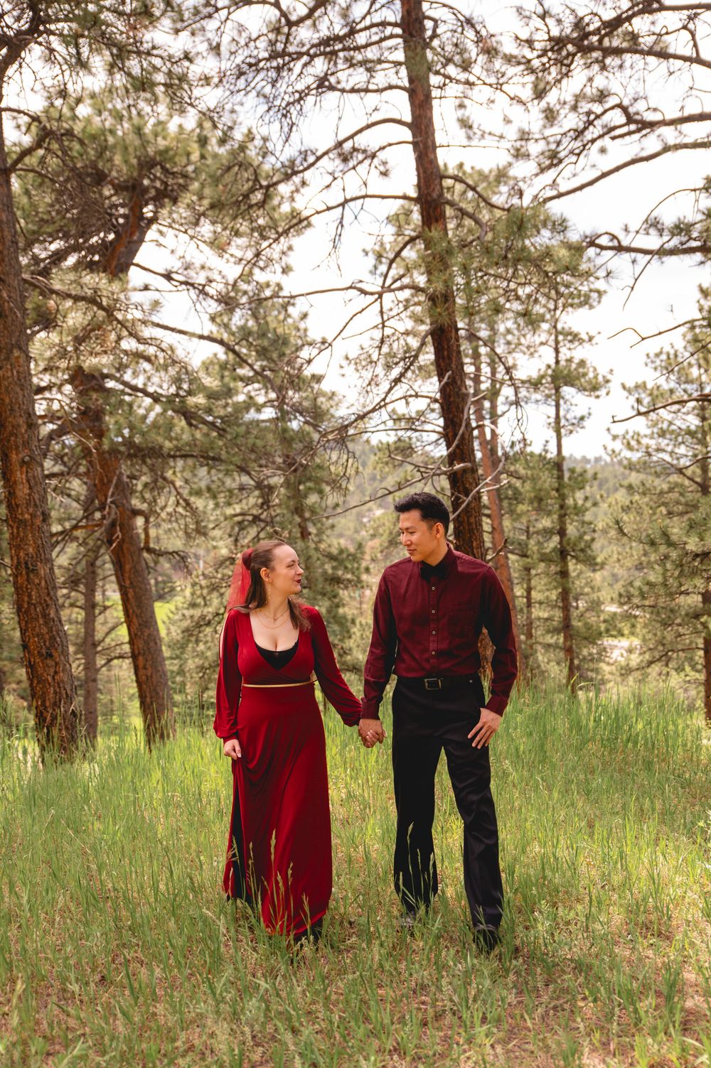 A couple in coordinated red and black outfits walks together through a serene pine forest setting.