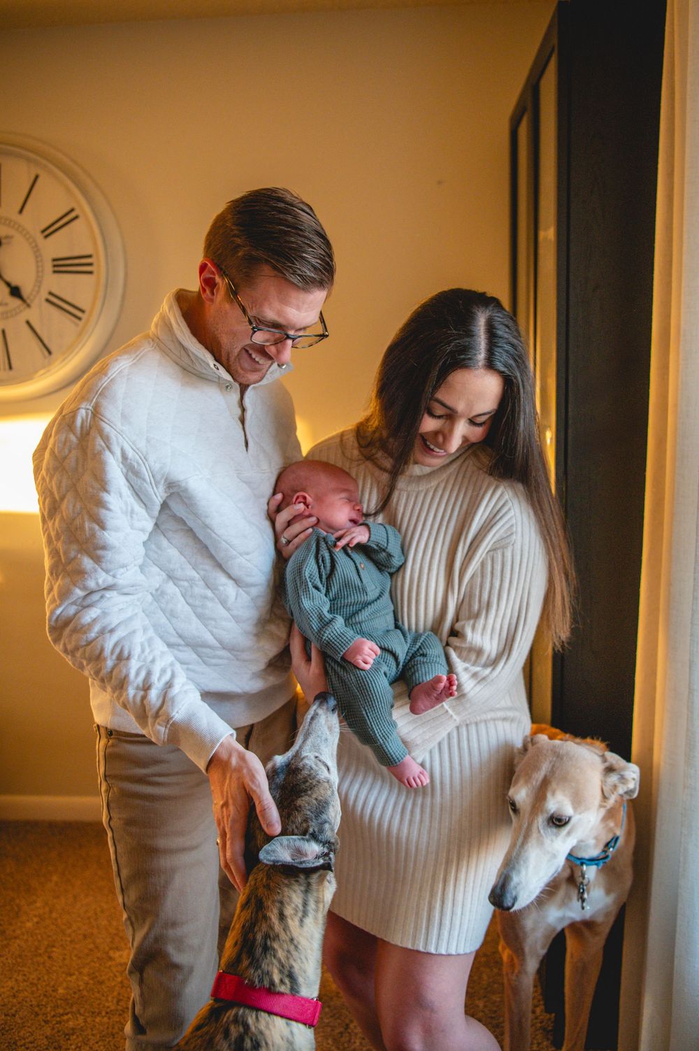 A couple in white and cream sweaters share tender moments with their newborn baby and dog in a warmly lit room.