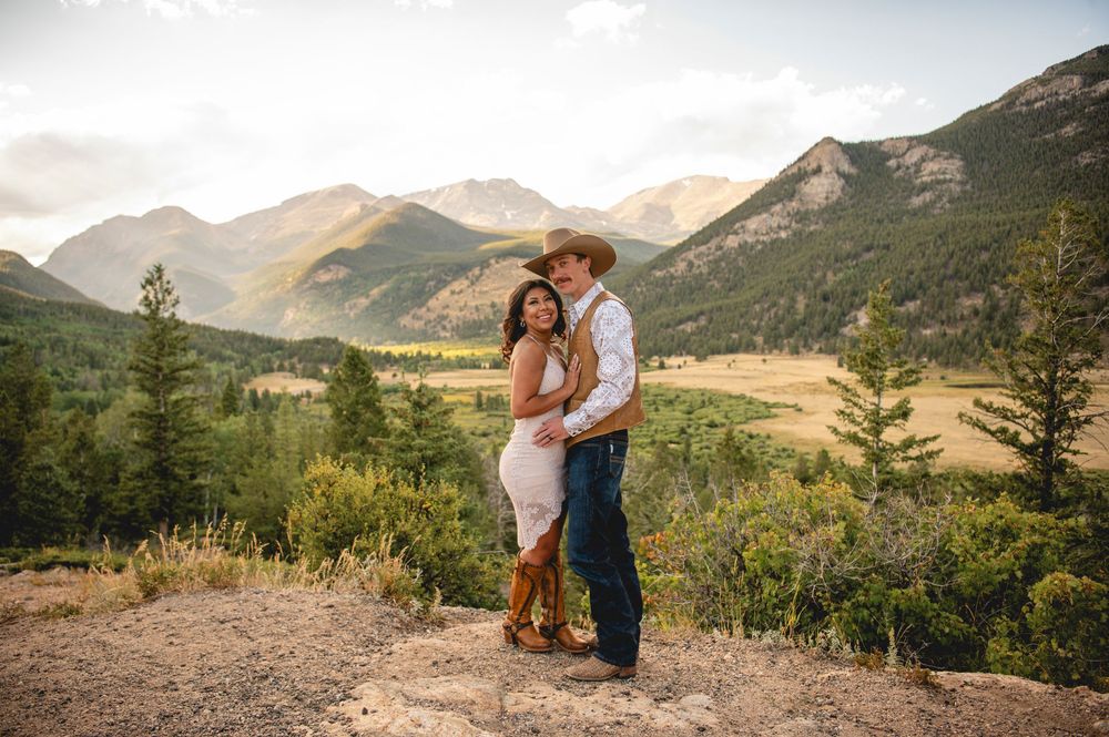 A couple embraces on a scenic mountain overlook with golden valleys and evergreen forests in the background.
