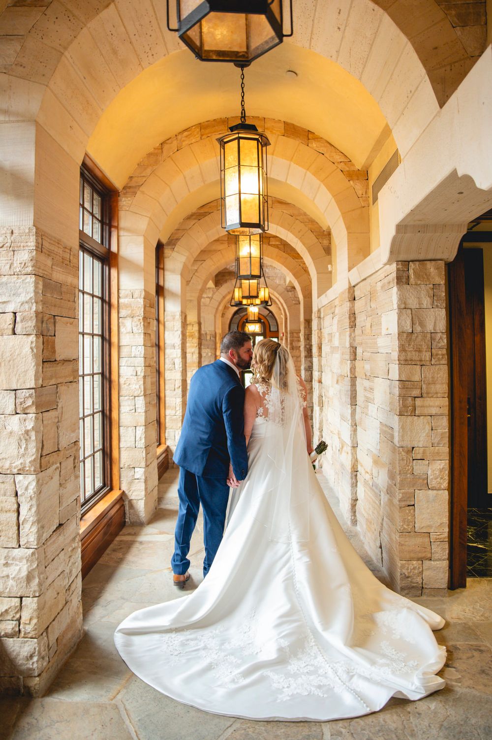 A couple walks through an elegant stone archway corridor with hanging lanterns and large windows casting warm light.