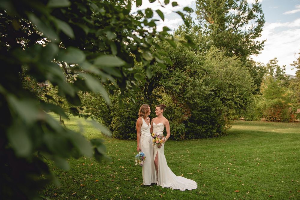 Couple in white dresses embrace in a lush green orchard during golden hour.