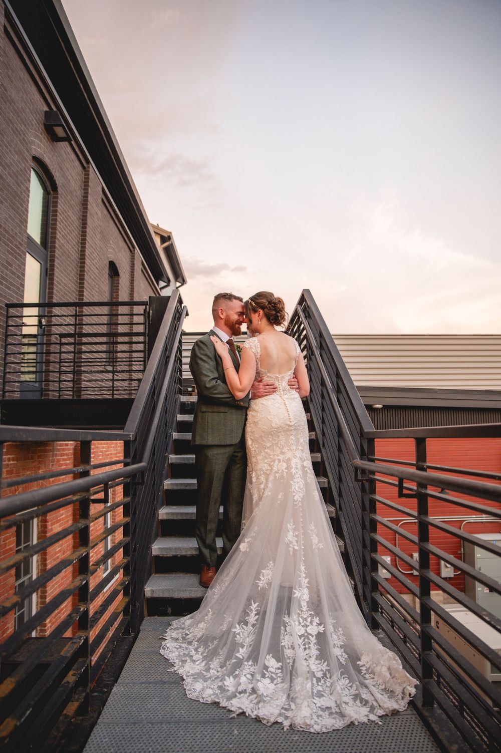 A romantic wedding couple embraces on a modern industrial staircase at sunset with a lace gown trailing down the steps.