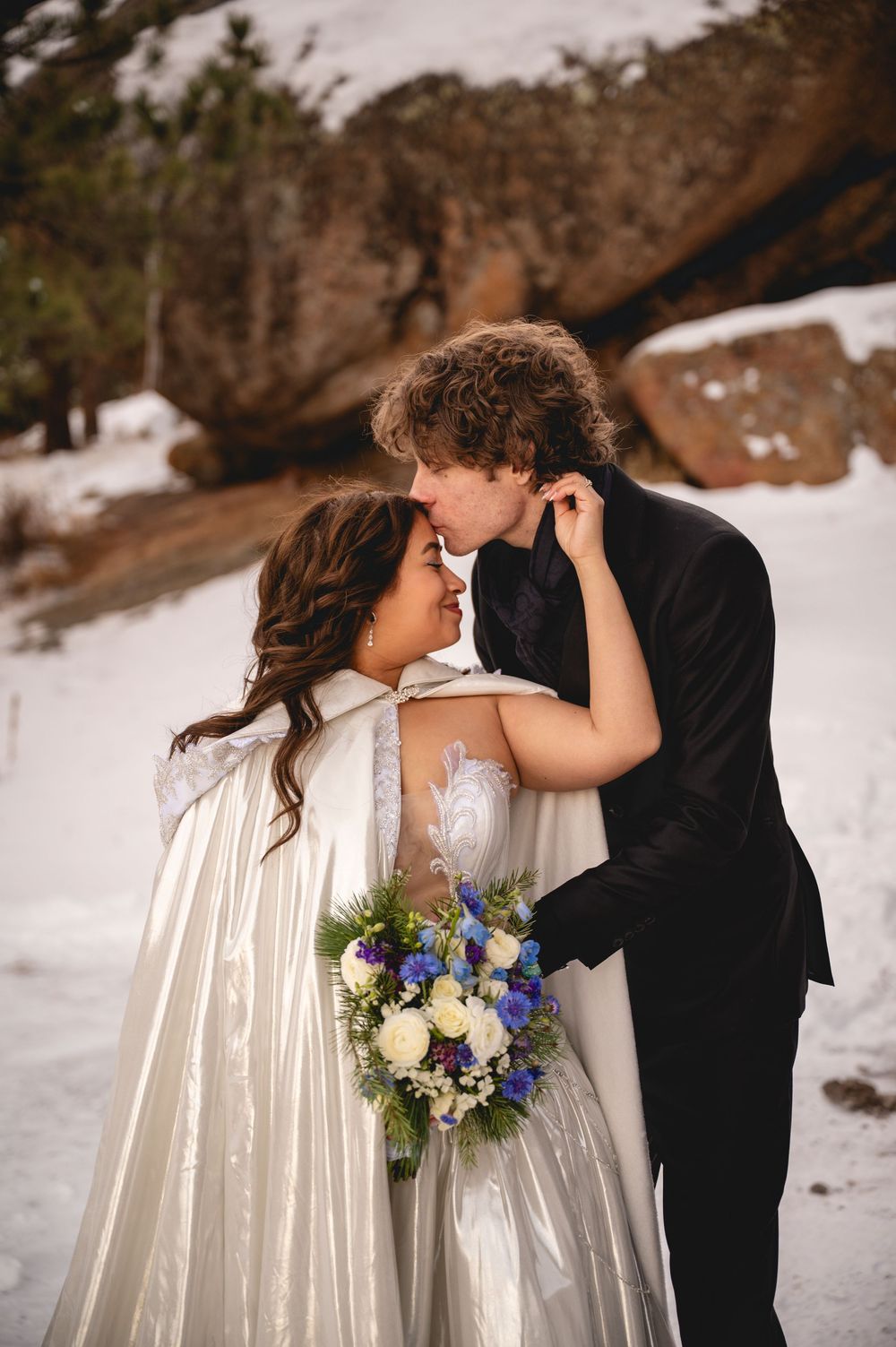 A couple shares romantic winter wedding moments in the snow with white and blue floral bouquet details.