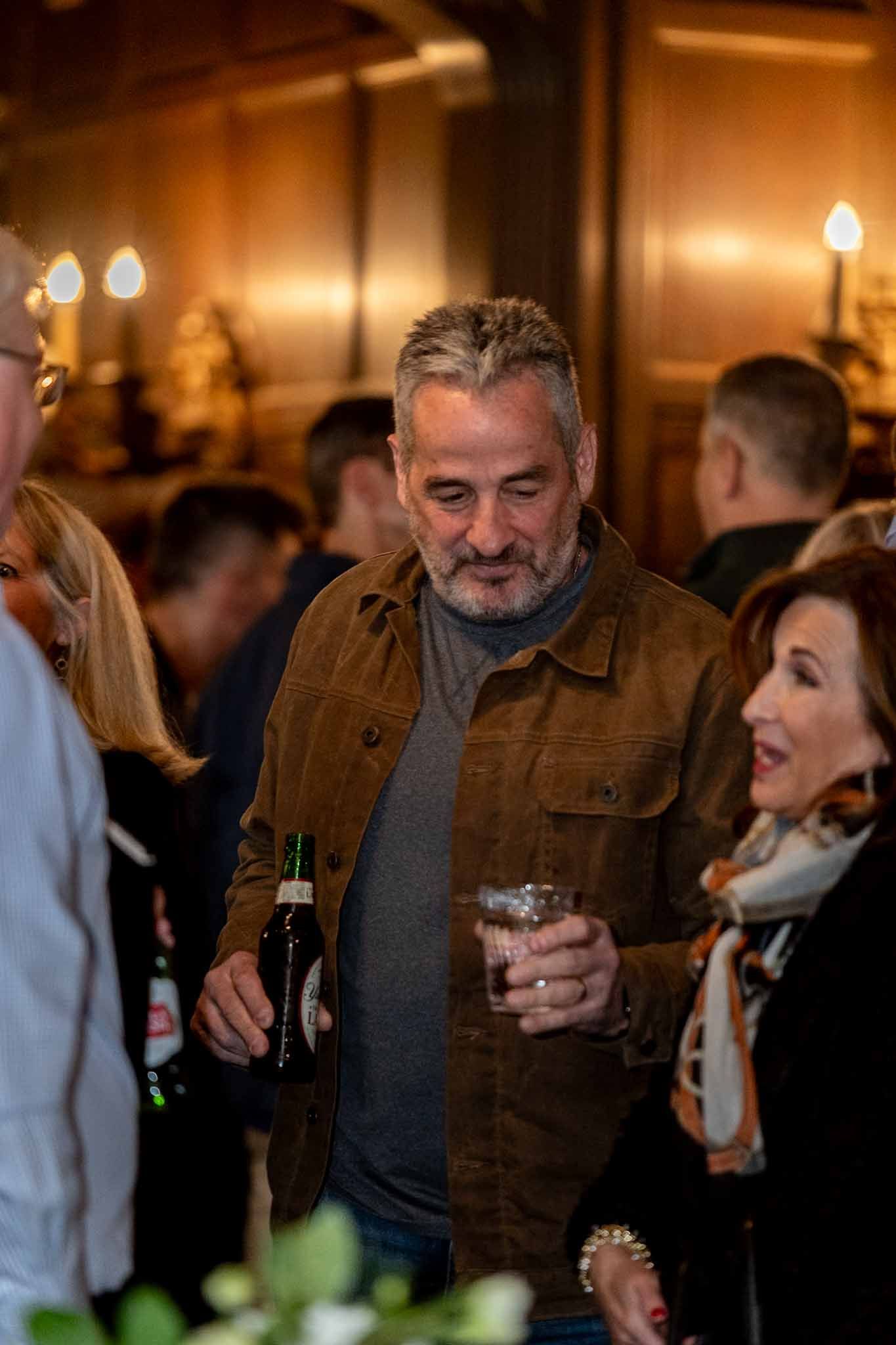 A person wearing a brown corduroy jacket holds a beer bottle and cocktail glass at an evening social gathering.