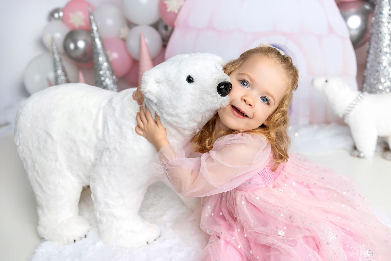 A young child in a pink tutu dress plays with white plush teddy bears and stuffed animals in a whimsical setting.
