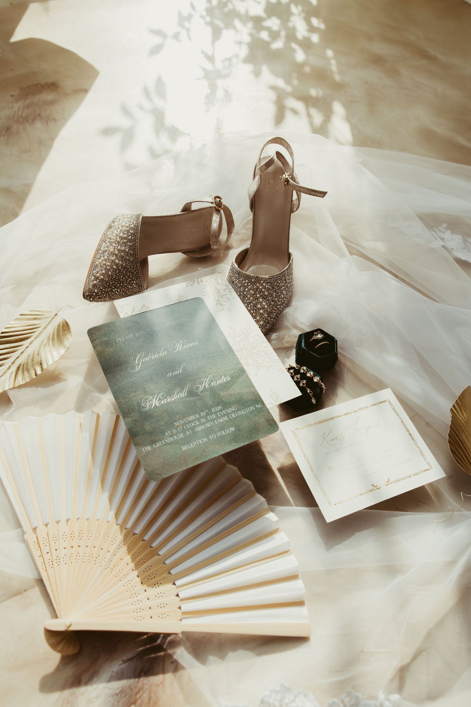 Elegant wedding flat lay with glittery heels, green invitation, hand fan, and ring box on veil.