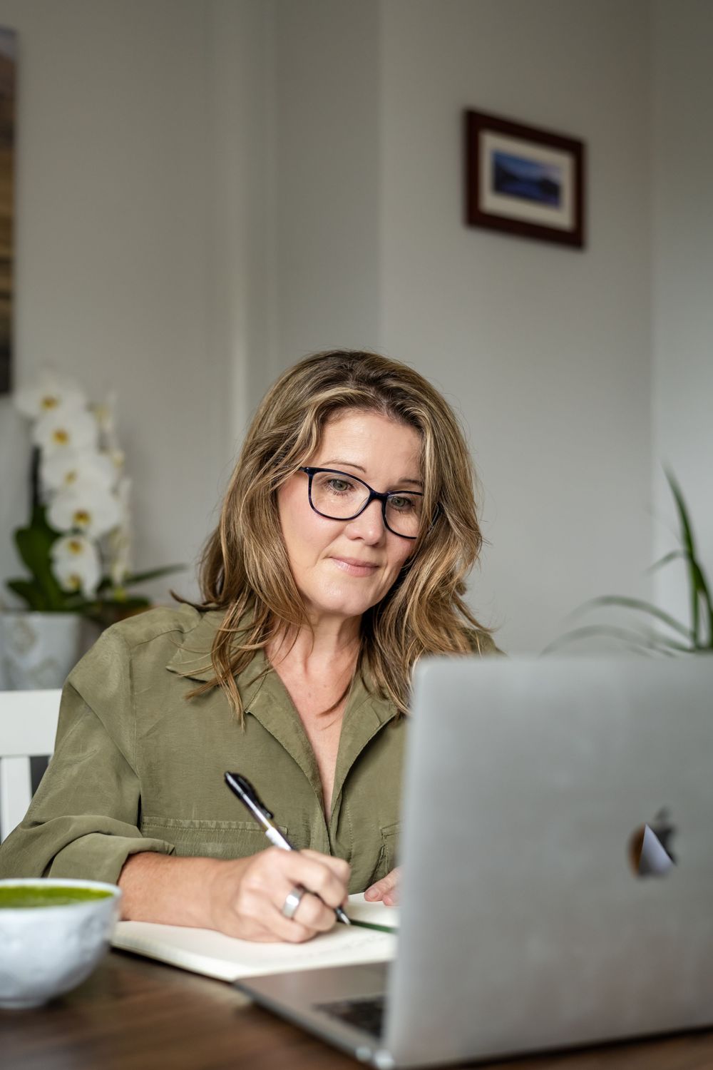 Professional wearing glasses writes in notebook at desk with laptop and orchid plant in background.