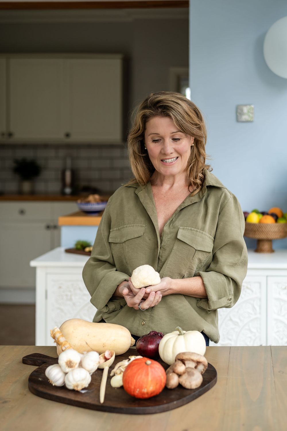 A person in an olive green shirt arranges fresh produce including mushrooms, apples, and bread on a wooden cutting board.