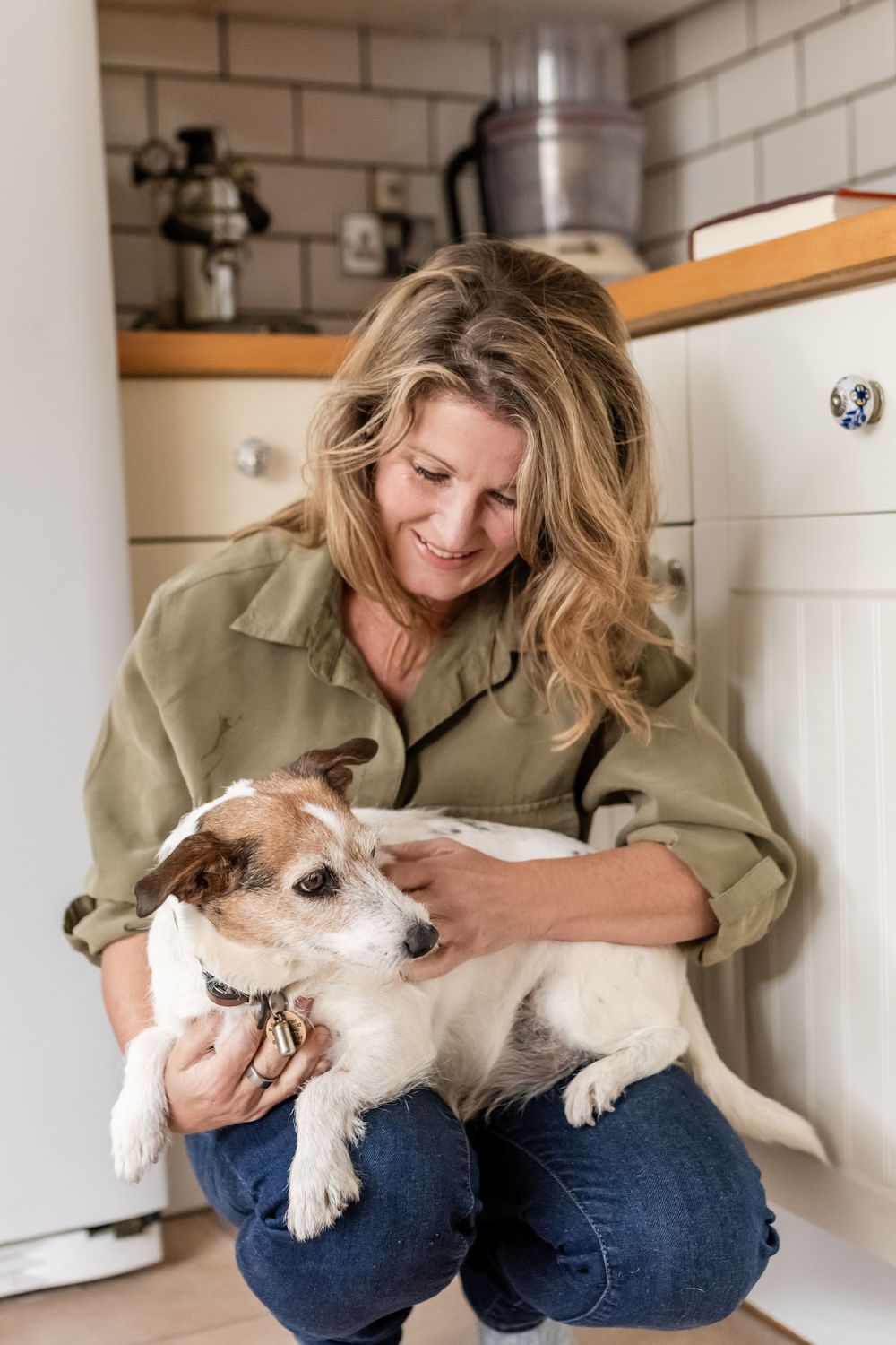 A person in jeans and olive shirt lovingly petting and interacting with a small white dog.