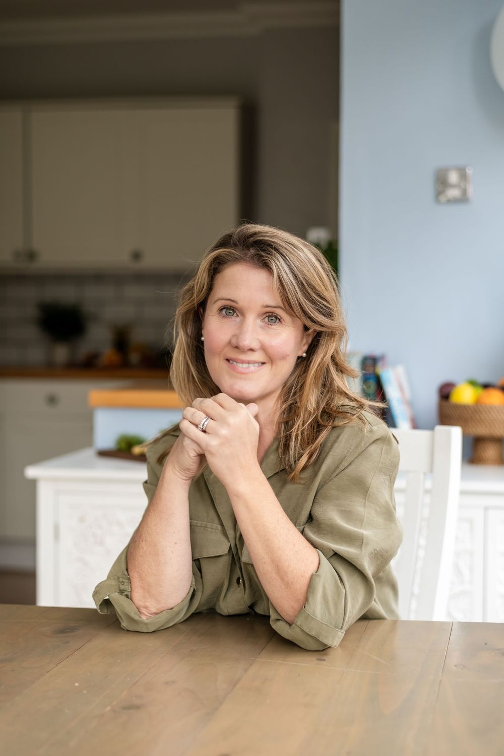Woman in olive green shirt rests head on hands while leaning on wooden table in bright kitchen setting.