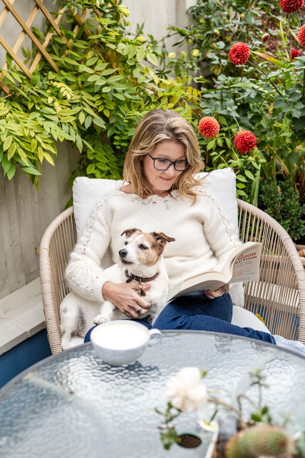 A woman reading a book with a small brown and white dog by her side on a patio chair surrounded by green plants and red flowers.
