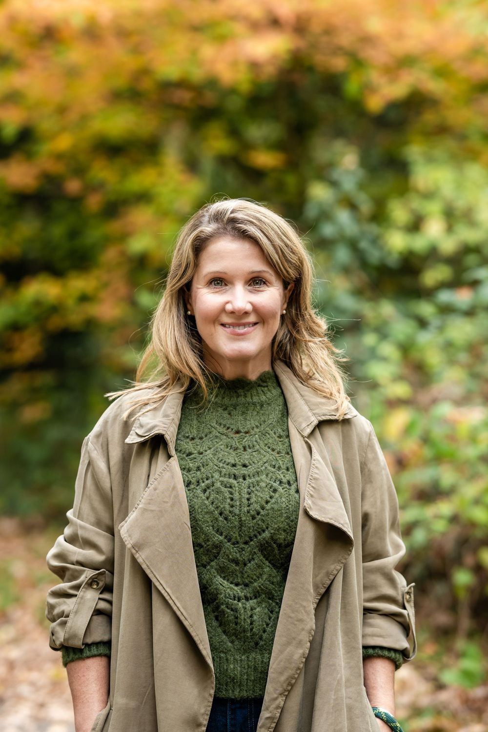 A person wearing a green knit sweater and tan jacket poses outdoors with autumn foliage in the background.
