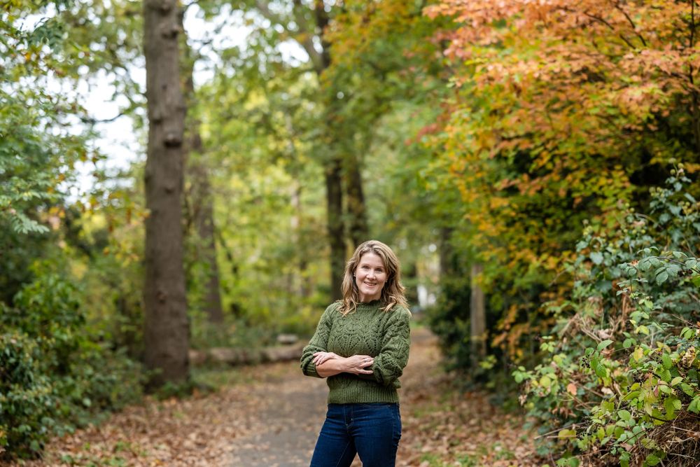 A woman stands in a scenic autumn forest setting with colorful fall foliage in the background.