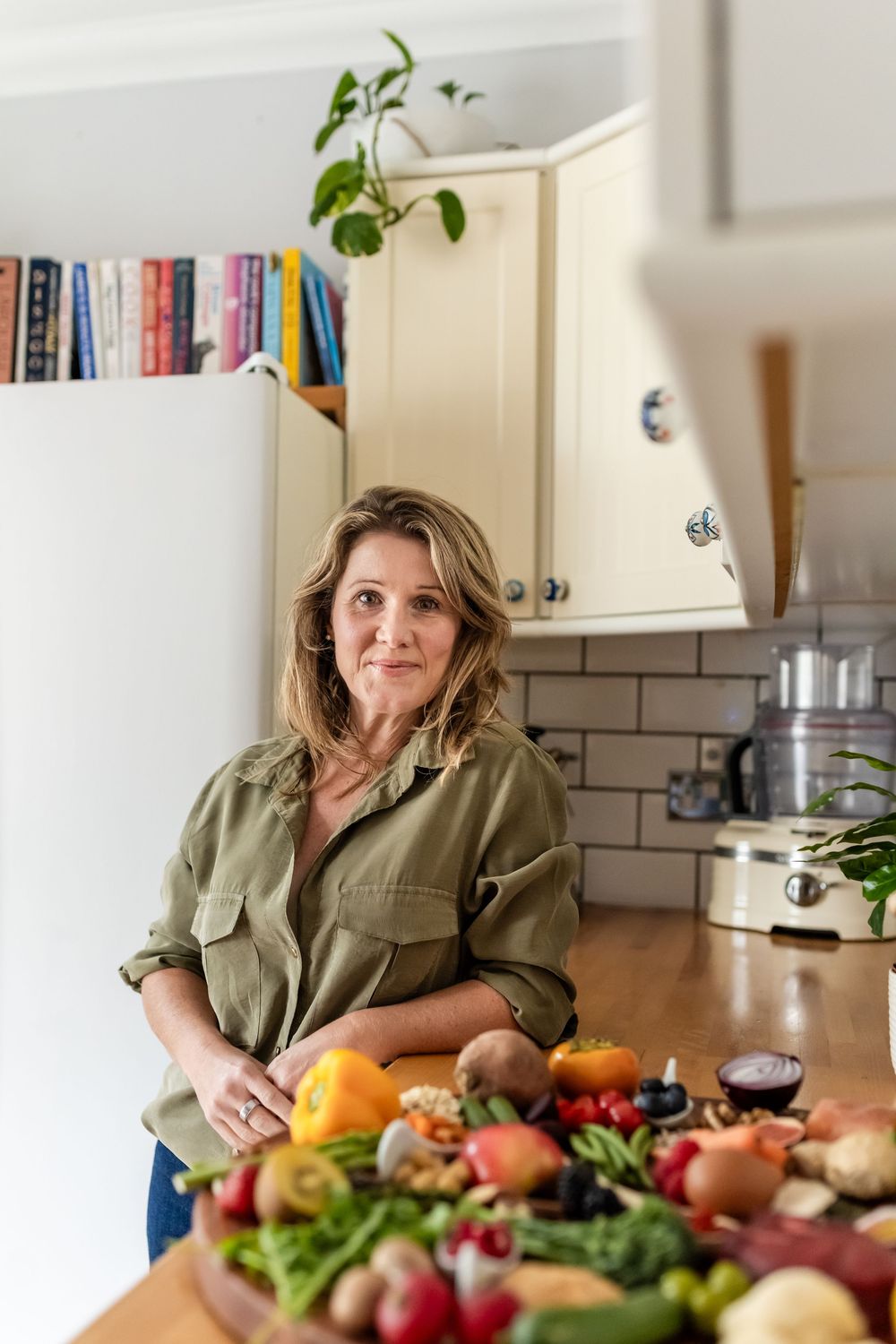 A woman stands in a bright kitchen with fresh produce and vegetables spread across a wooden countertop.