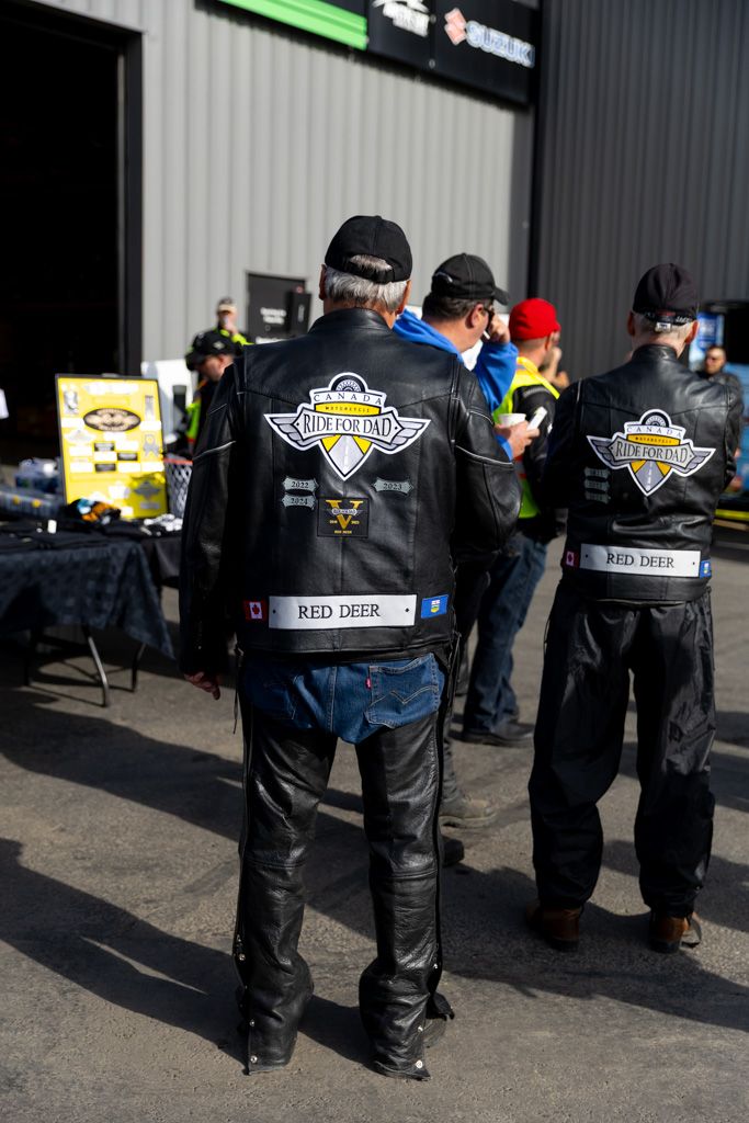 Group of motorcycle club members wearing leather jackets with patches standing outside in a row.