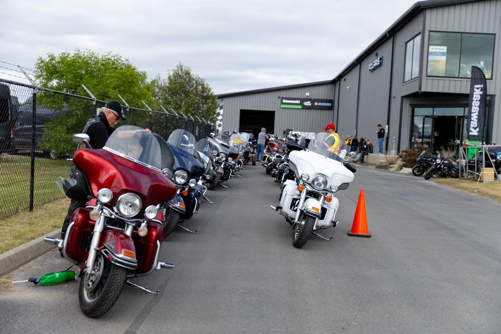 Row of motorcycles parked outside a gray industrial building with riders gathering for an event.
