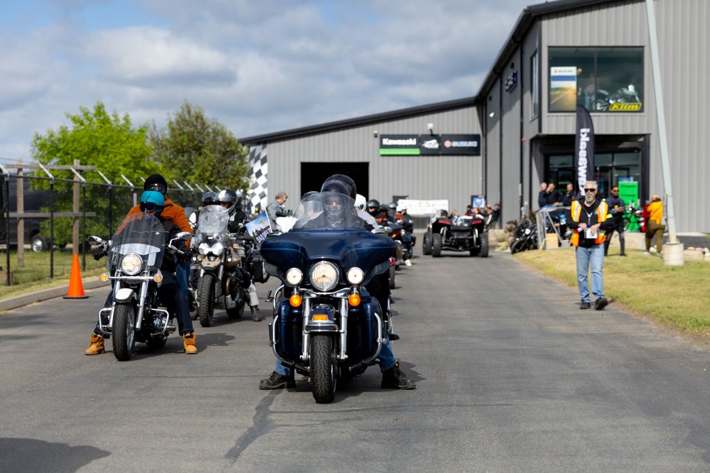 Row of motorcycles parked outside a large industrial building with riders gathered in the background.