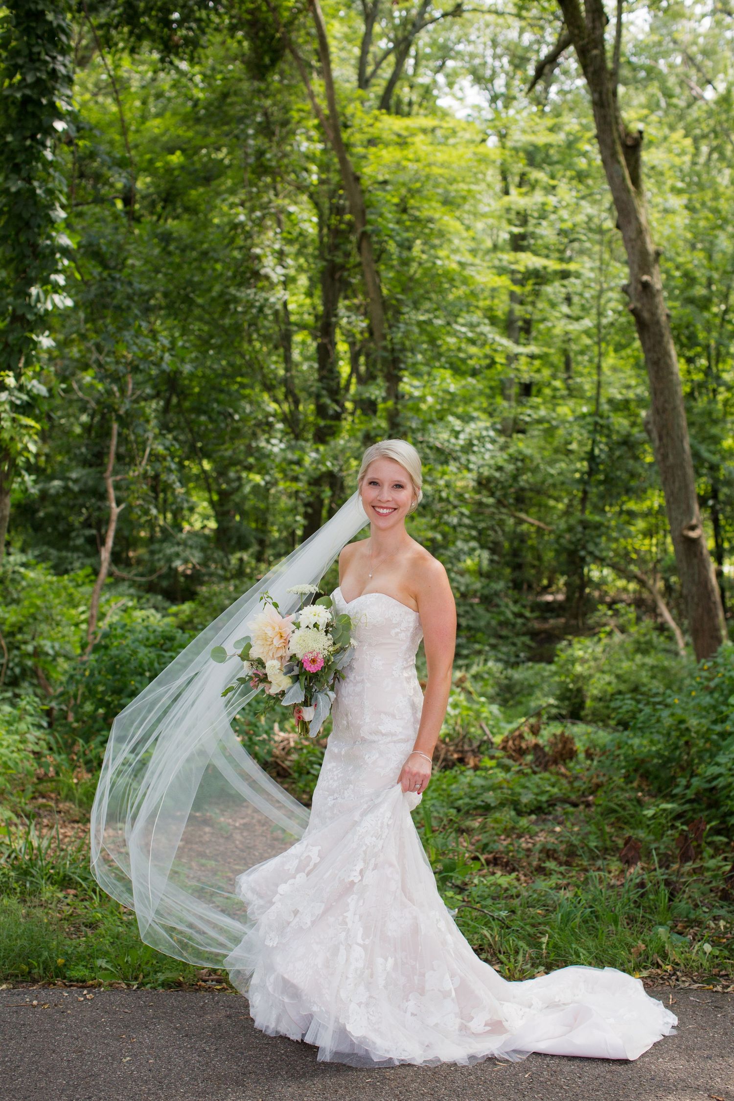 A bride in a lace wedding gown poses outdoors in a lush green garden setting.