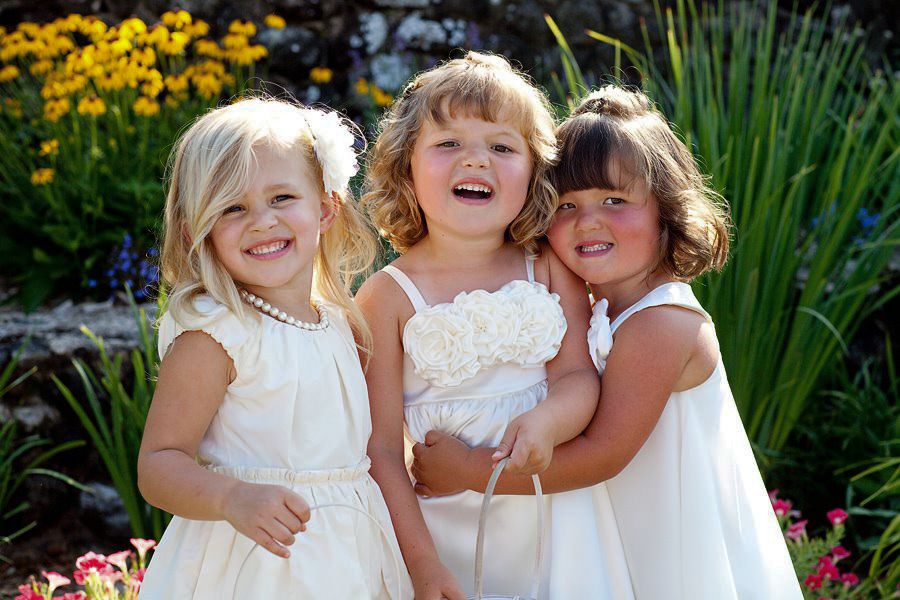 Young flower girls in white dresses pose together in a garden with yellow flowers and greenery.