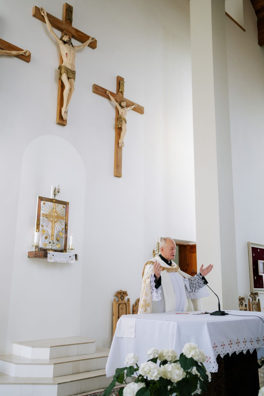 A priest performing a Catholic wedding ceremony beneath crucifixes mounted on a white church wall.