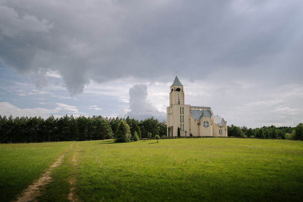 A white church with a tall steeple stands on a green lawn under dramatic cloudy skies.