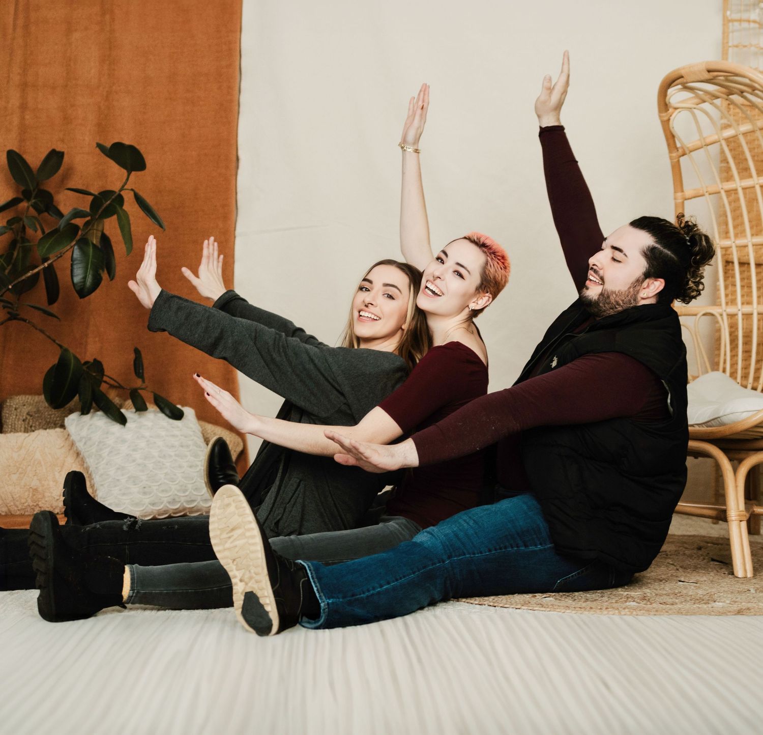 Three friends lounging on floor with raised arms and joyful expressions in a cozy home setting with rattan chair.