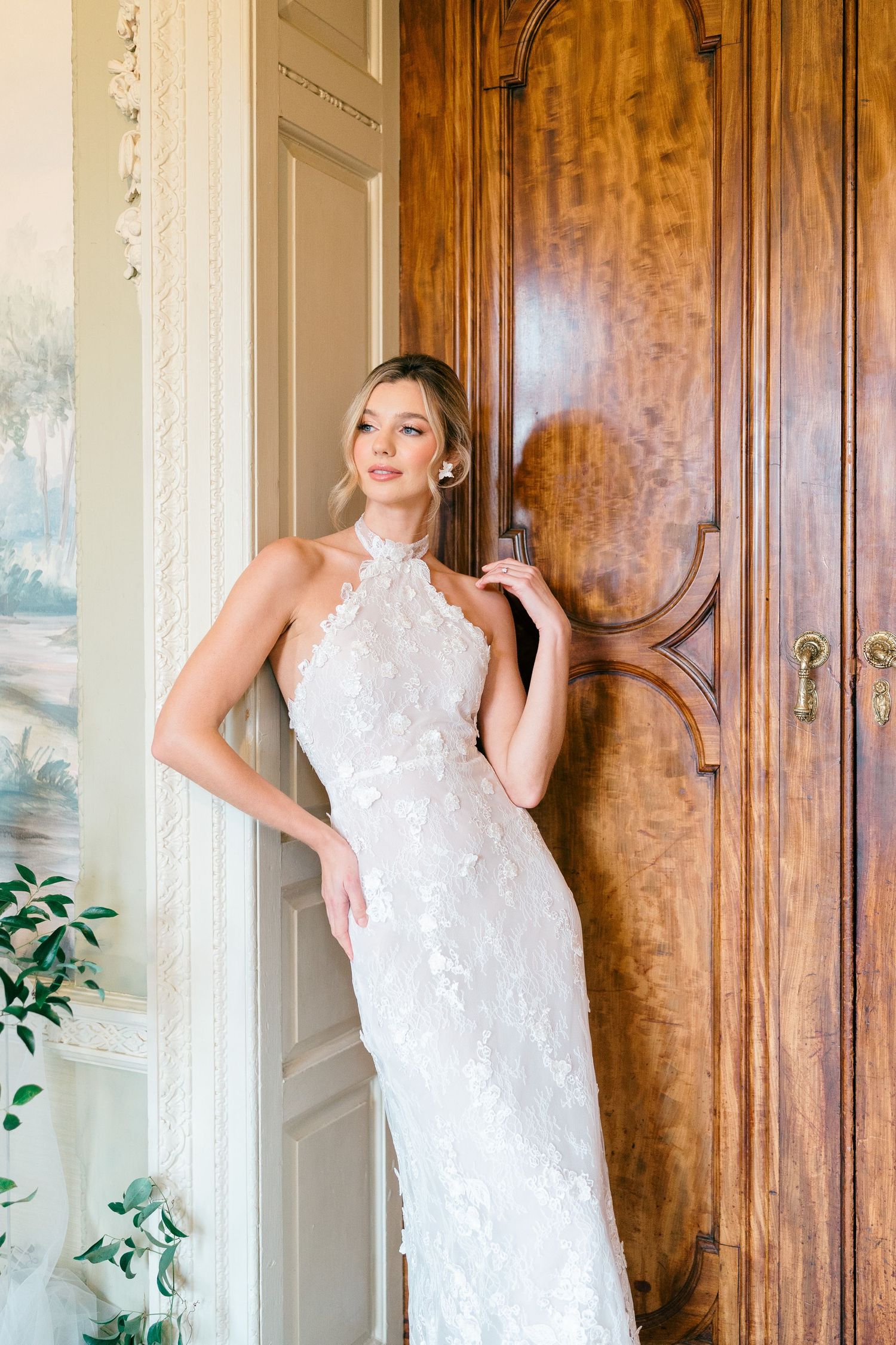 A bride in an elegant white lace gown poses gracefully against a wooden paneled wall with greenery accents.