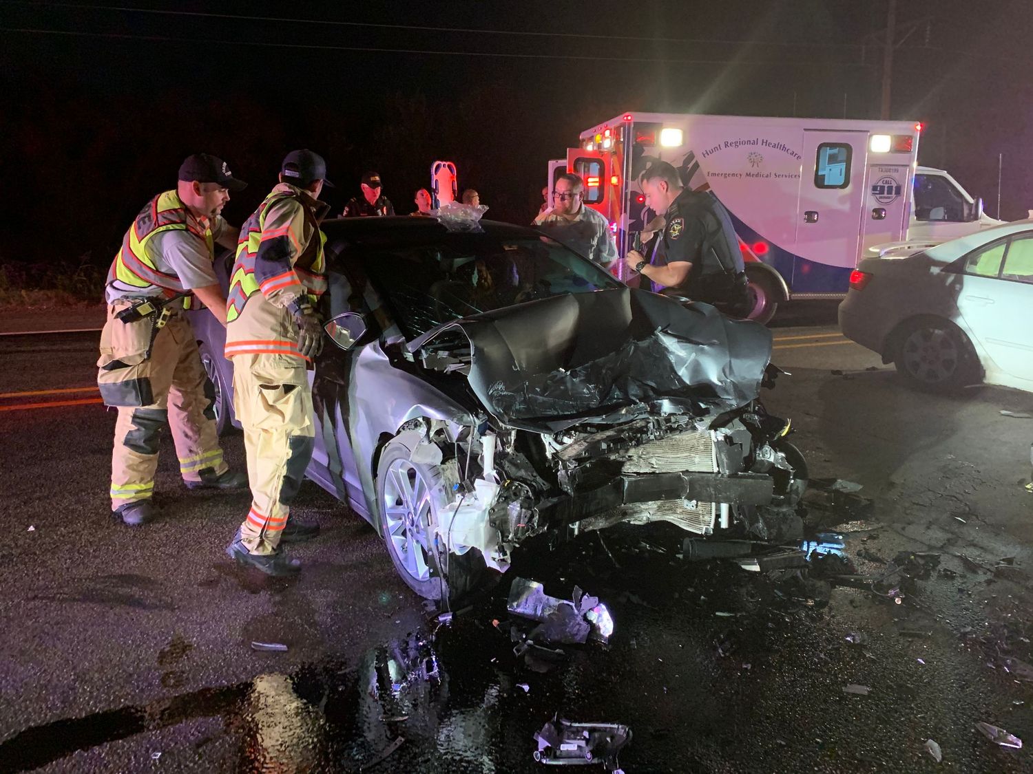 Emergency responders examine a damaged vehicle at night with an ambulance visible in the background.