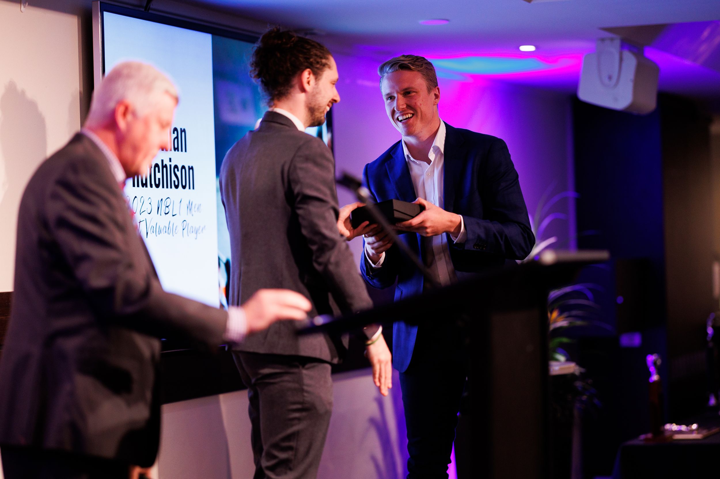 Three people engaged in conversation at a business event with purple lighting and presentation screens.