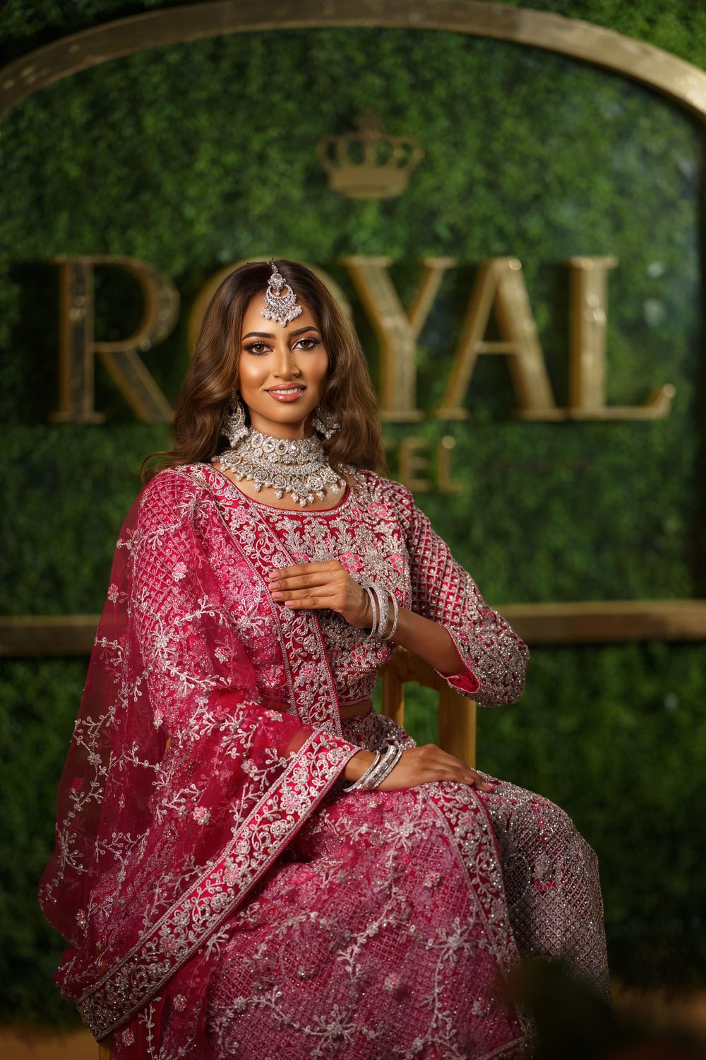 Traditional Indian bridal attire in deep pink with silver embellishments shown against a green backdrop with royal crown decor.