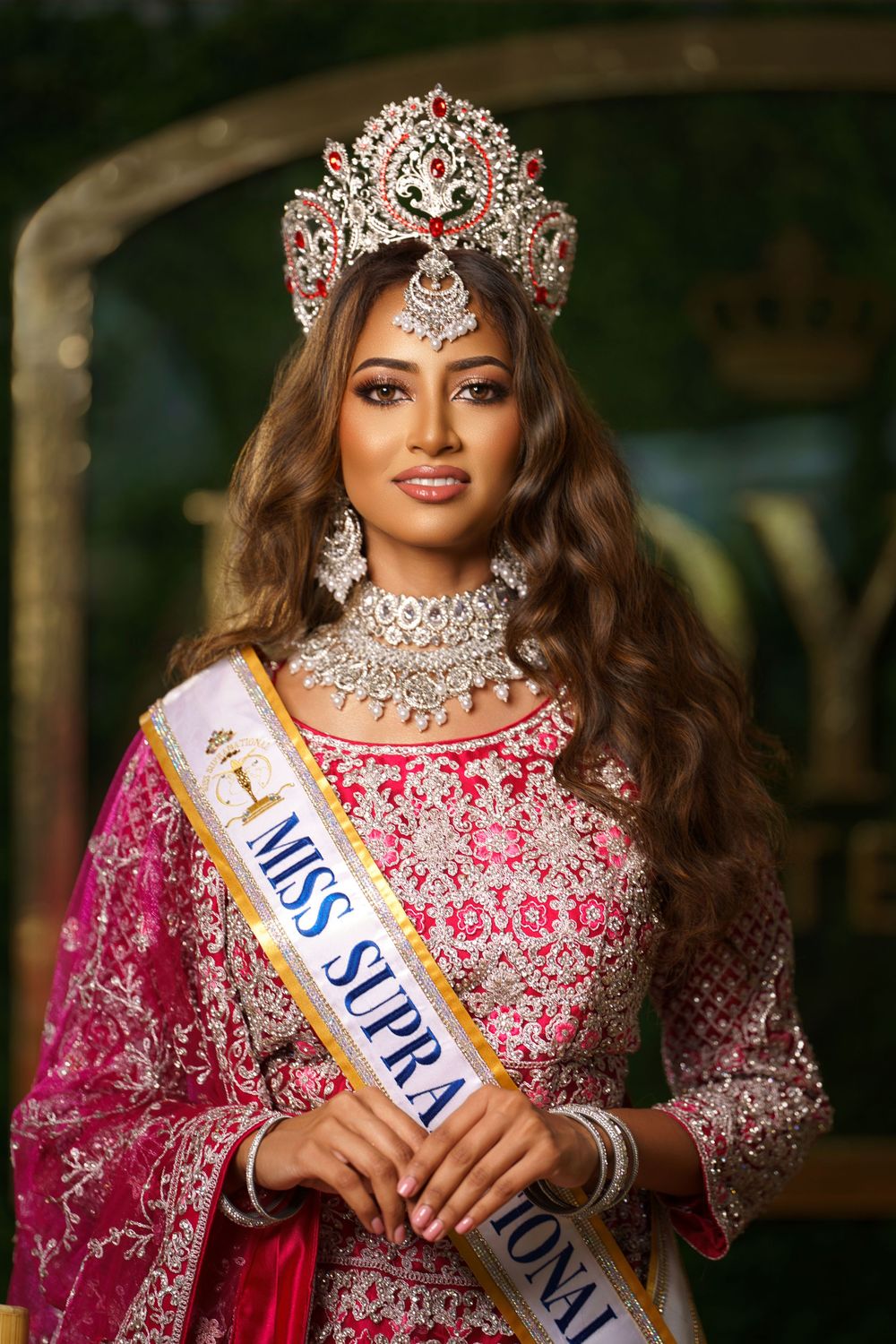 Beauty pageant contestant wearing ornate crystal crown and pink embellished traditional Indian lehenga with jewelry.