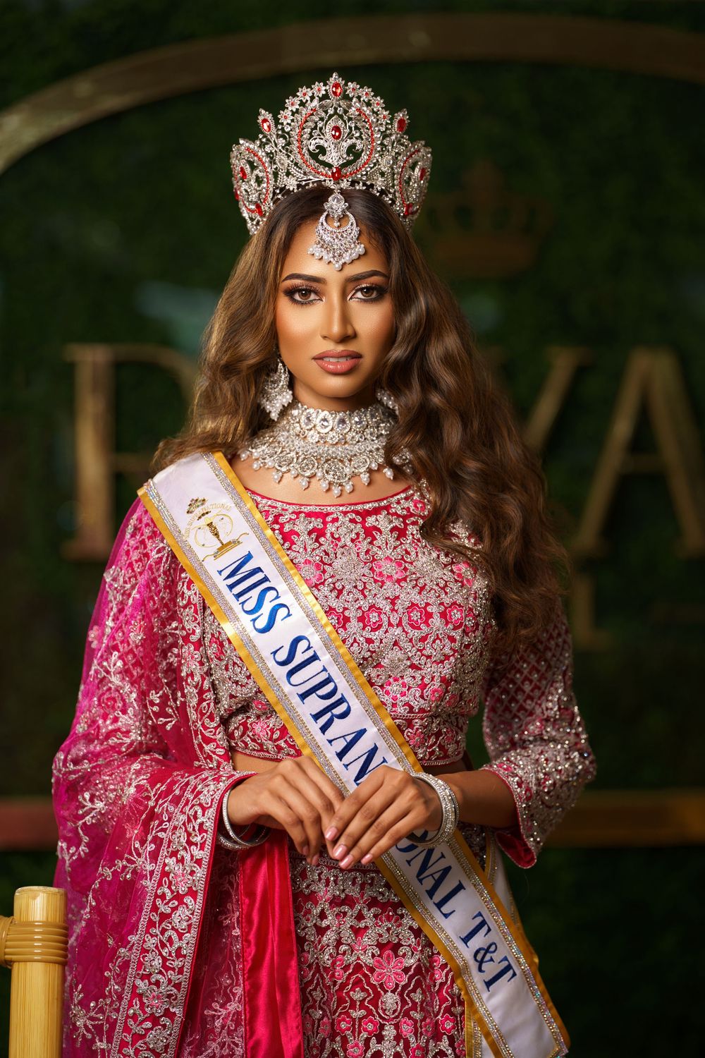 Beauty pageant contestant wearing ornate crown and pink embellished traditional dress with Miss Supranational sash.