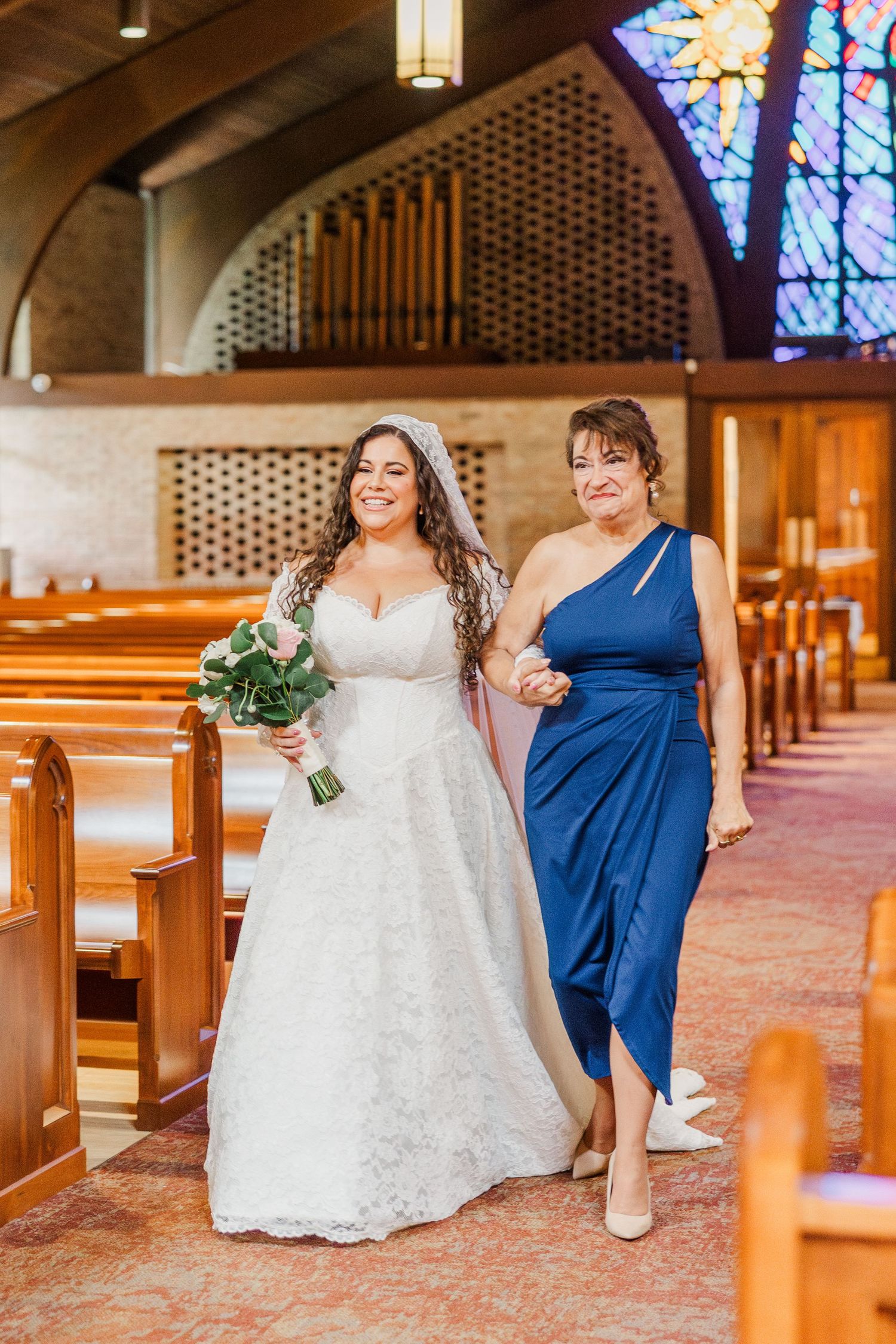 Wedding party members in white and navy blue dresses pose playfully in a church aisle with stained glass windows.