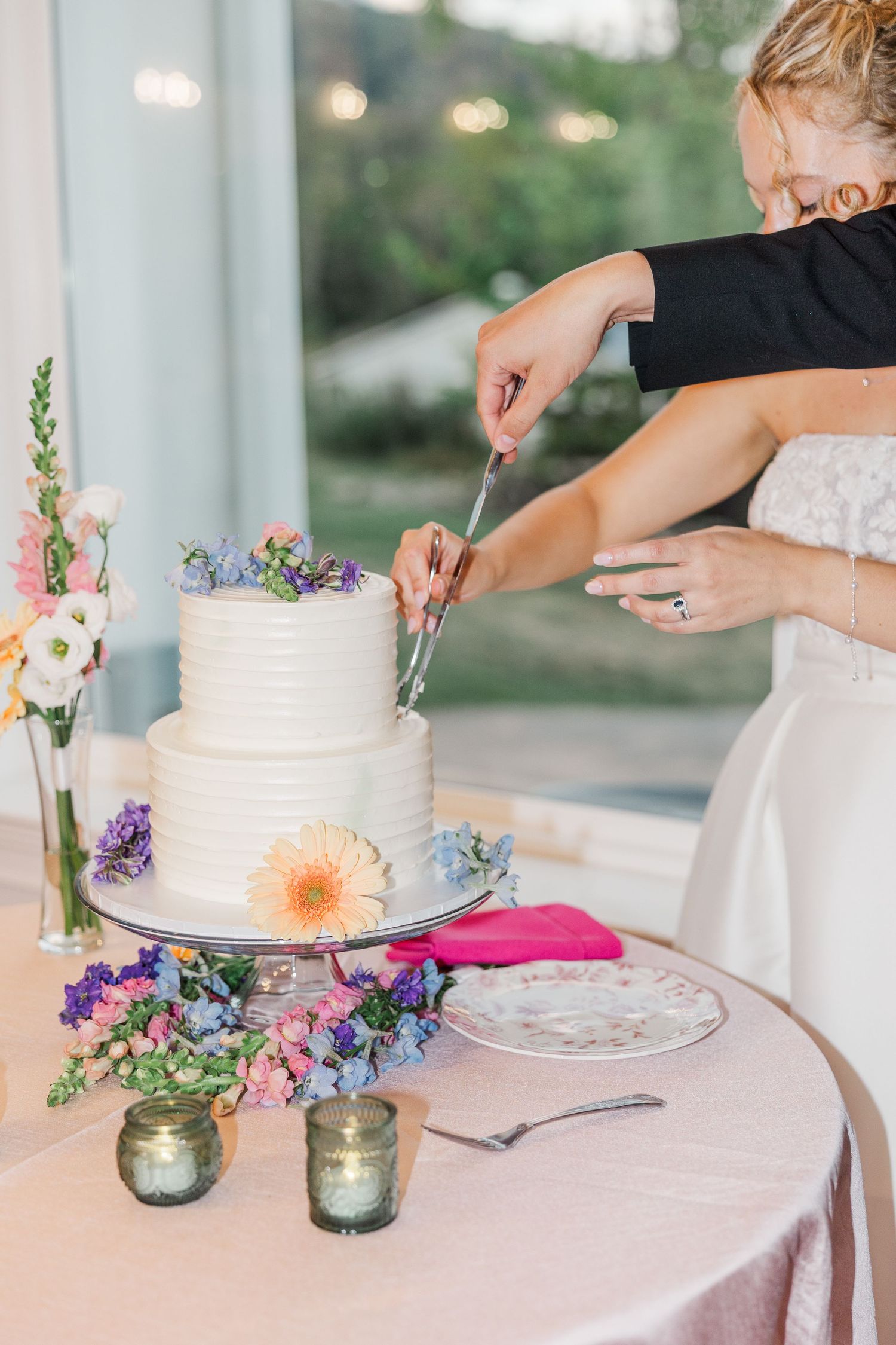 A white wedding cake with delicate floral decorations being cut on a table with mason jar candles.
