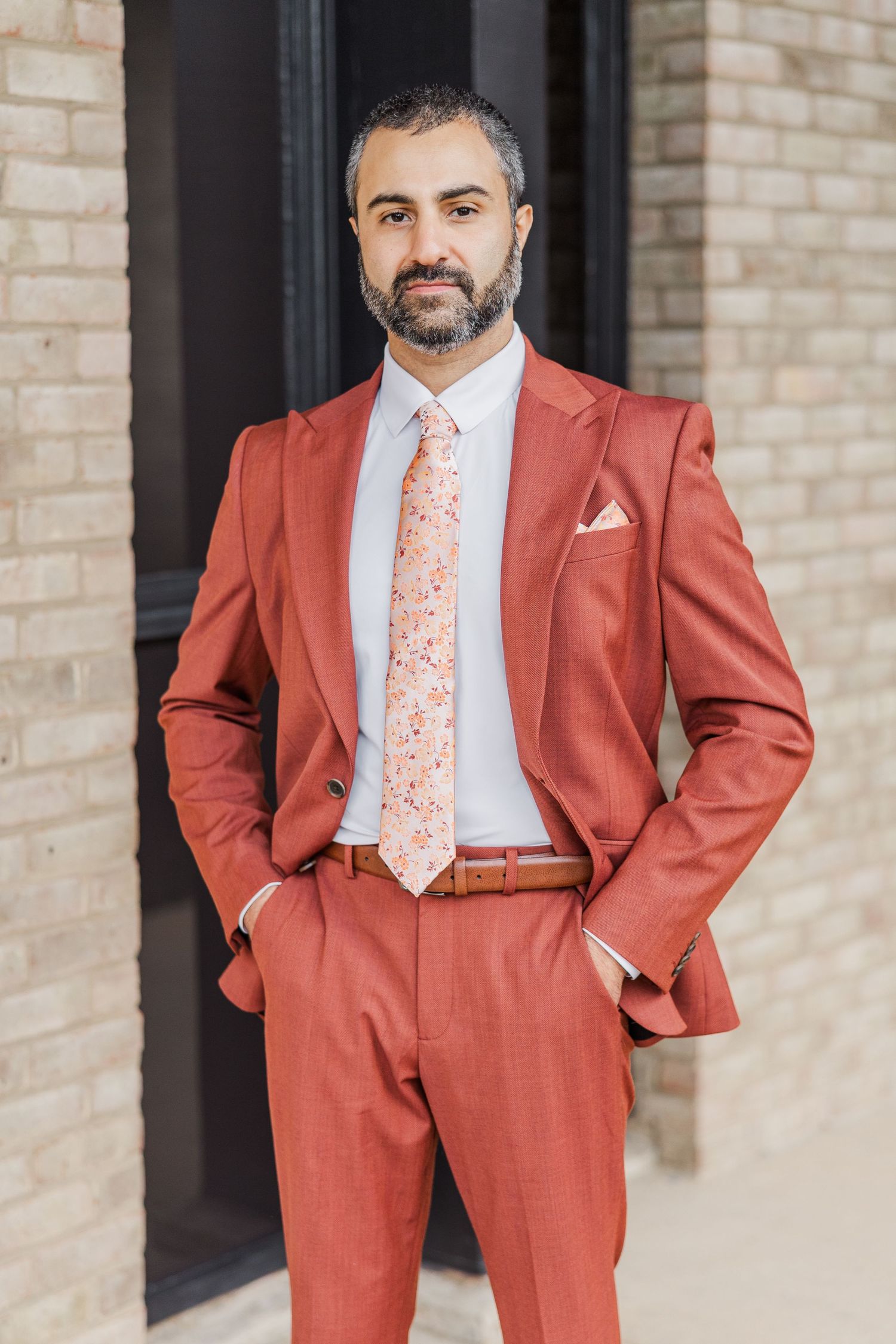 Professional wearing a stylish terracotta suit with floral tie and white dress shirt poses against brick wall.