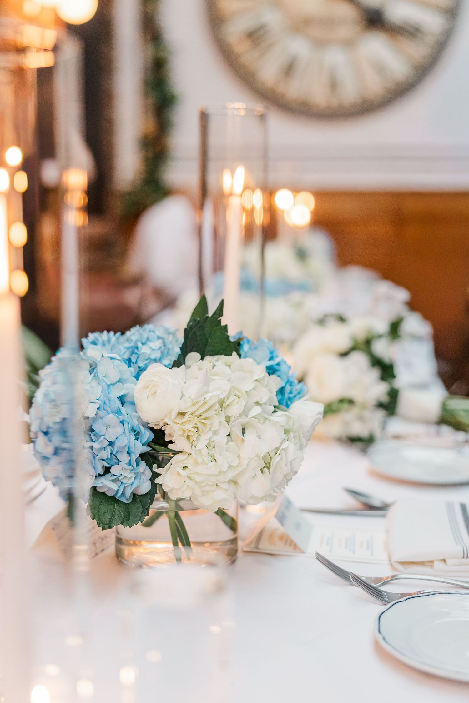 Elegant wedding table setting with blue and white hydrangea centerpieces and glowing candlelight in the background.