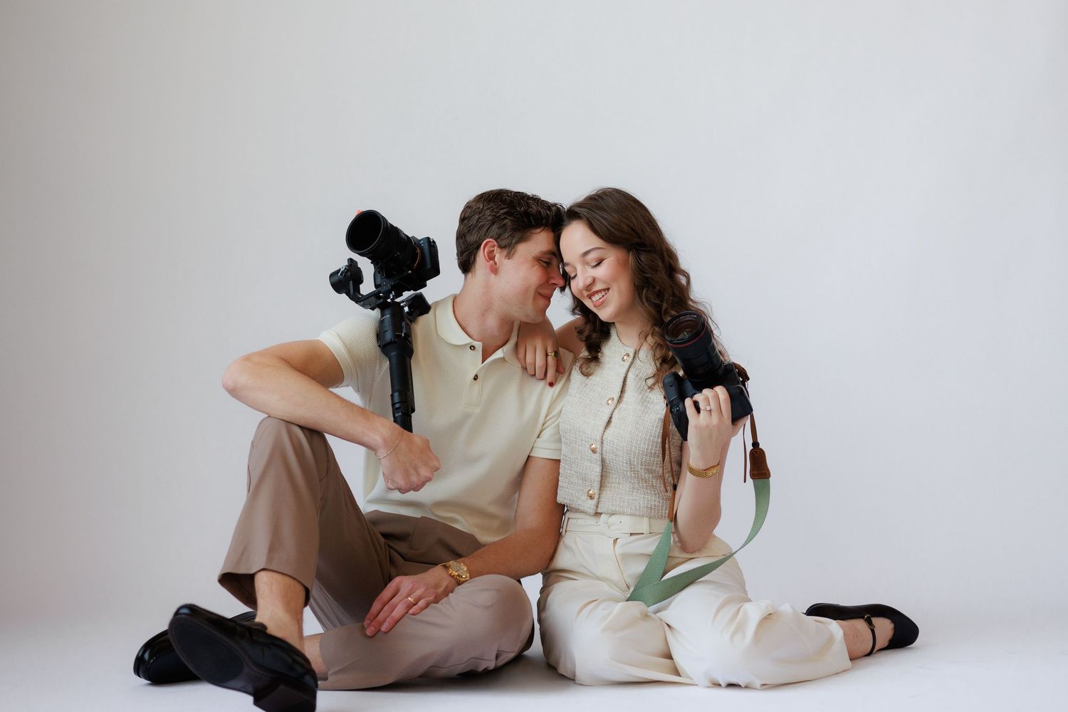 A couple in casual attire poses together near a window in a warm-lit indoor setting.