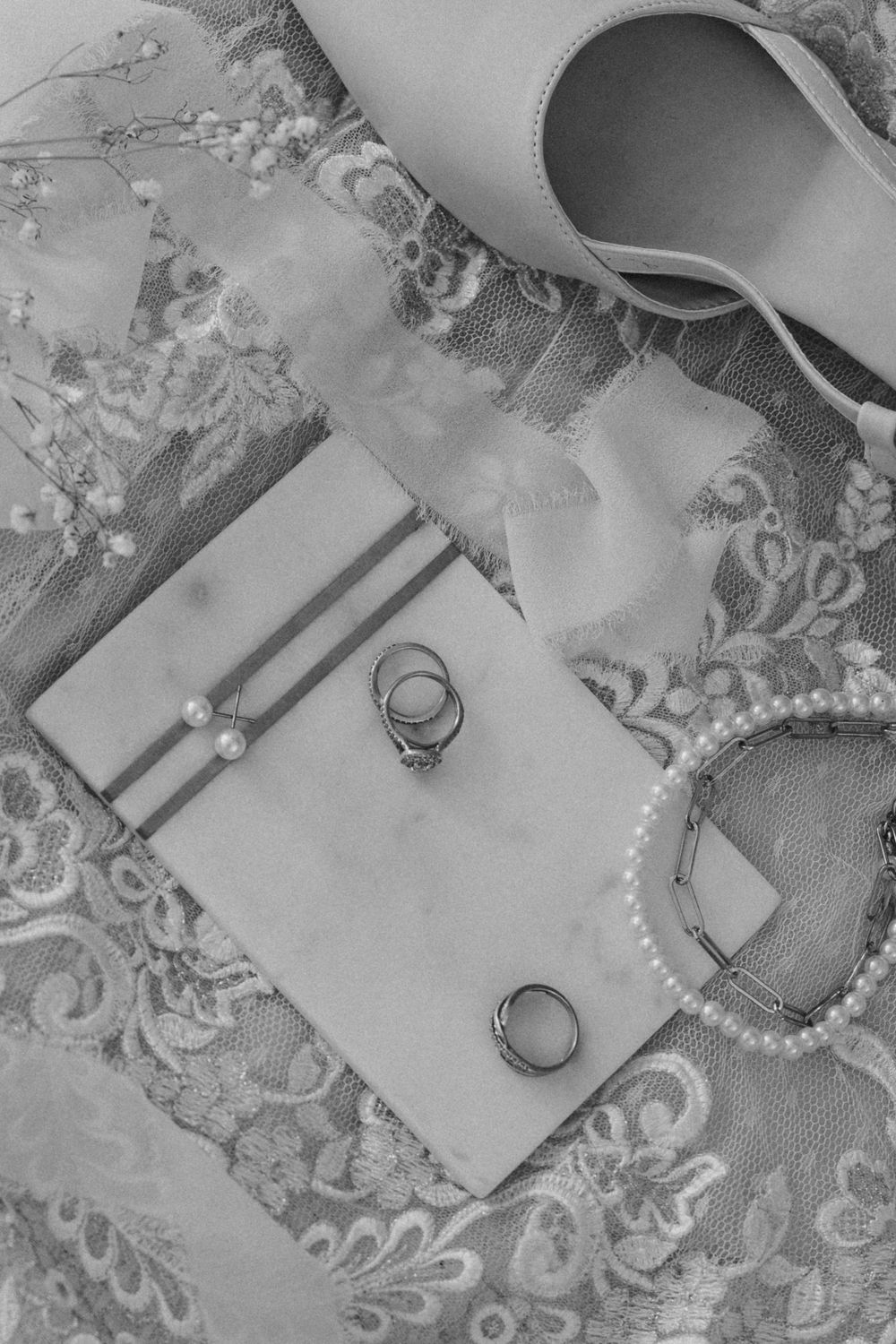 Wedding rings and bridal accessories arranged on delicate lace fabric in black and white photography.