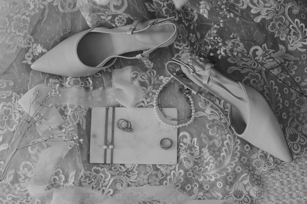 Elegant white bridal shoes and accessories arranged on a delicate lace background with floral patterns.