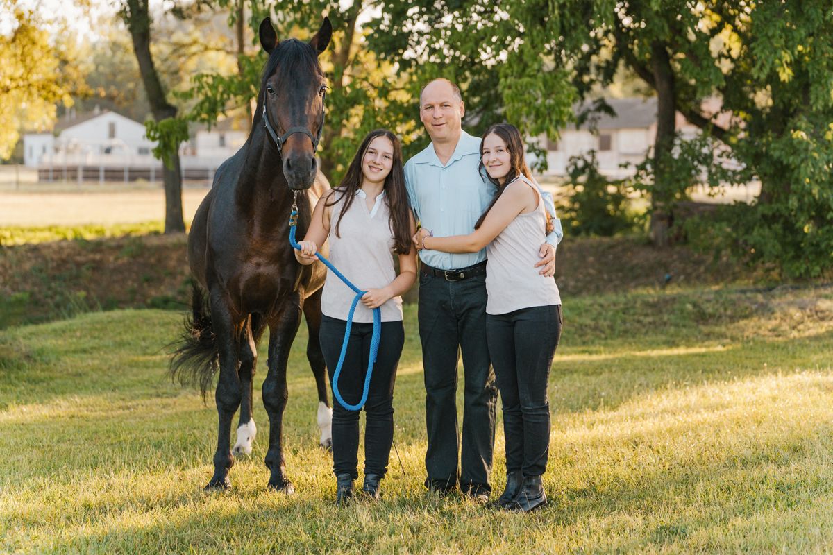 A beautiful black horse stands with dad and his daughters on a grassy field during golden hour family photoshoot in the hills district
