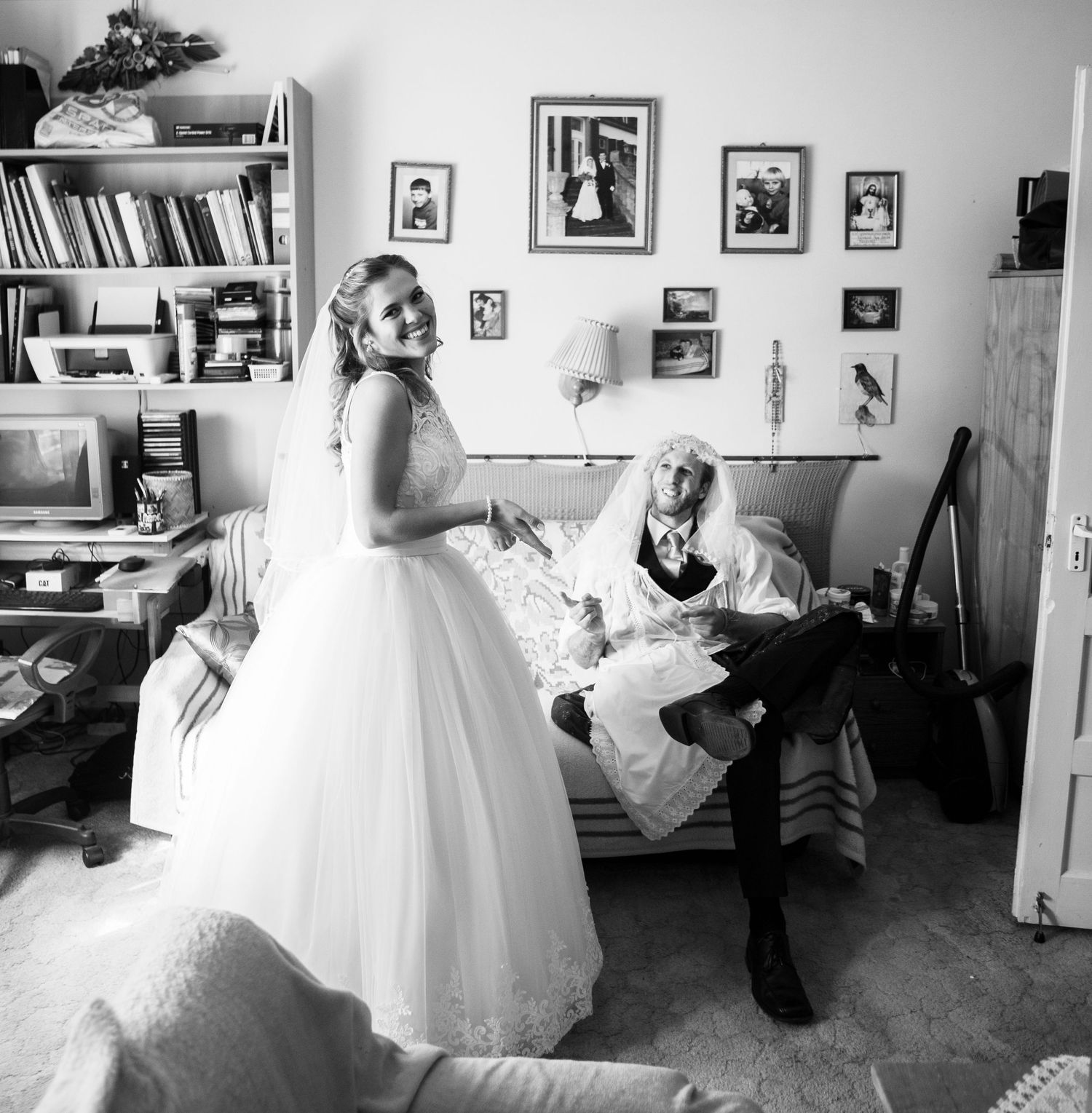 Black and white photo showing a person in a flowing white wedding dress standing near someone relaxed on a bed in a book-filled room.