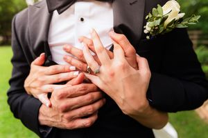 Newlywed couple displaying wedding rings, groom in black tuxedo with boutonniere, bride embracing him from behind outdoors. DFW Wedding