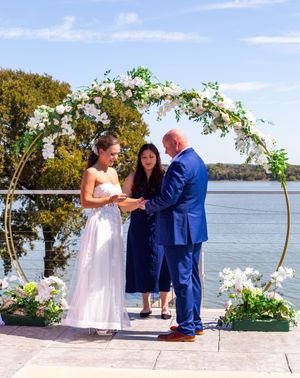 A wedding ceremony takes place under a floral arch by a serene lakeside with white flowers and greenery. Eagle Mountain Fort Worth wedding
