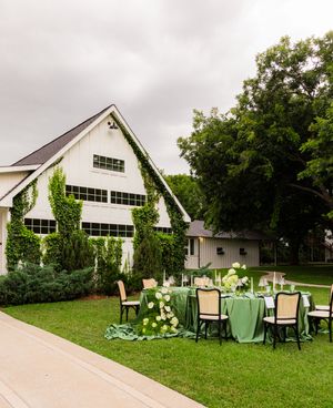 Elegant outdoor wedding reception setup on green lawn beside ivy-covered white barn venue under overcast skies.